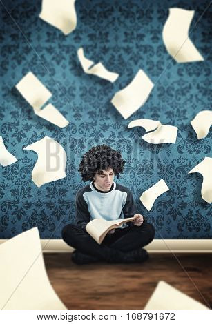 Young guy reading a book with flying sheets coming out of the book in a room.