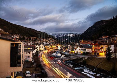 Val Gardena, Italy. Evening on the popular winter resort Ortisei of Val Gardena, Italy. Dolomites alps mountains at sunset. Illuminated hotels, cafes and restaurants, car traffic lights