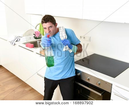 young attractive and happy man holding washing detergent spray bottle and cloth wearing rubber gloves smiling confident as aiming with hand gun playful enjoying housework and domestic duty