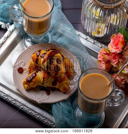 Croissants drizzled with chocolate lying on a tray. Two transparent cups with coffee nearby. On the right are the flowers and candle