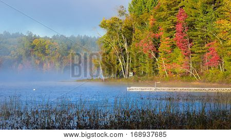 Late-summer, morning sun shines on mist rising from warmer wooland lake water.  Dock extends from lakeside cottage country in a deciduous waterfront forest.