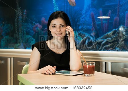 Stylish brunette woman sitting at a cafe drinking juice and talking on a cellphone in the background blue aquarium