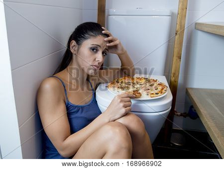 young sad and depressed bulimic woman feeling sick guilty sitting at the floor of the toilet leaning on WC eating pizza in nutrition disorder bulimia concept
