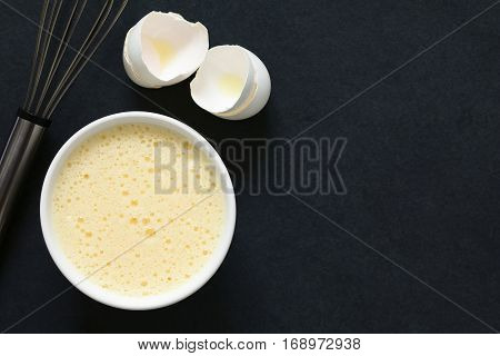 Whisked whole eggs (whites and yolks together) in bowl with whisk and egg shells on the side photographed overhead on slate with natural light