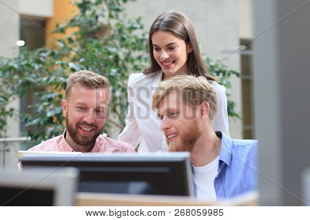 Group Of Young People In Casual Wear Sitting At The Office Desk And Discussing Something While Looki