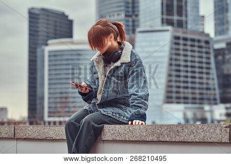 A Redhead Hipster Girl With Tattoo On Her Face Using A Smartphone Sitting In Front Of Skyscrapers In