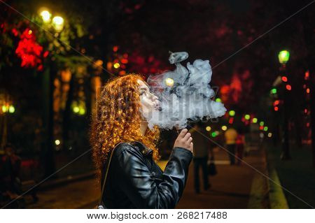 Young Red-haired Girl Smokes An Electronic Cigarette. She Walks Alone On Streets Of Night City. Non-