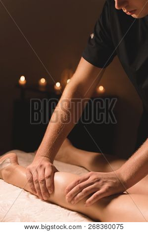 Close-up Of Male Hands Doing Calf Massage Of Female Legs In A Dark Room With Candles In The Backgrou