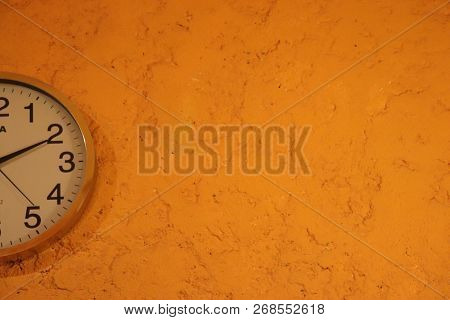 Round Wall Clok. The Background Of The Wall Has A Rough Texture And It Appears Orange,