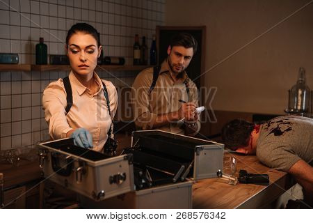 Concentated Female And Male Detectives Using Evidence Collection Kit And Investigating Dead Body At 