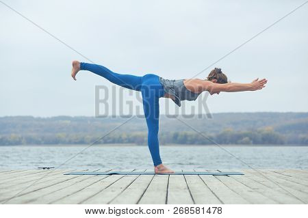 Beautiful Young Woman Practices Yoga Asana Virabhadrasana 3 - Warrior Pose 3 On The Wooden Deck Near