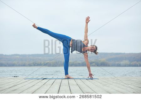 Beautiful Young Woman Practices Yoga Asana Ardha Chandrasana - Half Moon Pose On The Wooden Deck Nea
