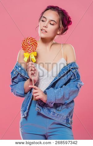 Pretty pink-haired girl with lollipop in denim jacket posing on a pink background. Fashion, beauty. Youth style of clothes.