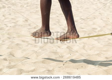 Closeup Man Feet Walking On Tightrope Or Slackline On Sandy Background. Slacklining Is A Practice In