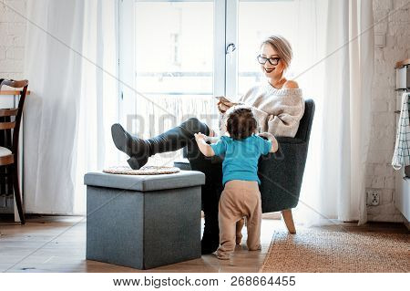 Young Mother With Mobile Phone Sitting In Armchair And Little Toddler Boy Stay Next To Her On A Fllo