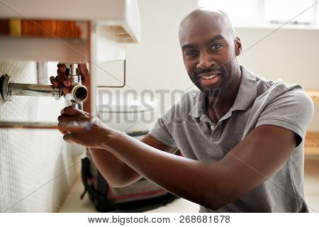 Young black male plumber sitting on the floor fixing a bathroom sink, looking to camera, close up