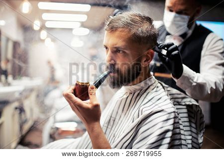 Hipster Man Client Visiting Haidresser And Hairstylist In Barber Shop, Smoking A Pipe.