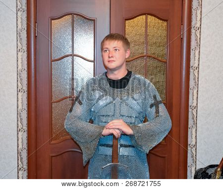 A Man Dressed Up In A Knights Hauberk At Home In His Room. Knights Sword In The Hands Of A Man. Knig