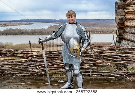Knight In Armor On The Background Of A Wicker Fence, A Wooden House And The Expanses Of The Motherla