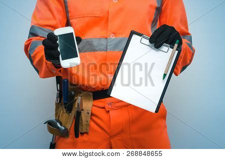 Building Contract. Fix List. Builder Is Holding Note Pad And Mobile Phone With Blank Screen In His H