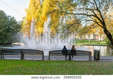 Two Unidentified Young Women Sit On A Green Painted Wooden Bench In The Park In Autumnal Colors. The