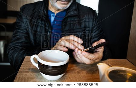 A Man Is Resting In A Cafe, Drinking Tea, Reading And Writing Messages On A Smartphone. Work In The 