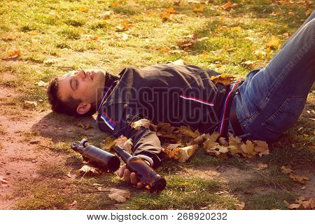 A Drunk Young Man Sleeping On Grass Under The Bench In The Day.a Young Man Holding A Bottle Of Alcoh