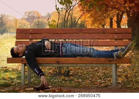 A Drunk Young Man Sleeping On A Bench In The Day.a Young Man Holding A Bottle Of Alcohol In Her Hand
