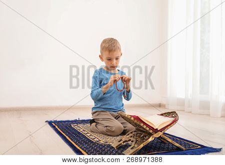 Little Muslim Boy With Misbaha And Koran Praying On Rug Indoors