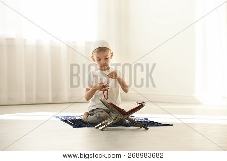Little Muslim Boy With Misbaha And Koran Praying On Rug Indoors