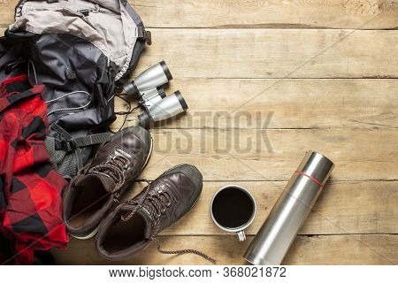 Hiking Boots, Binoculars, Shirt, Thermos, Backpack On A Wooden Background. The Concept Of Hiking, To