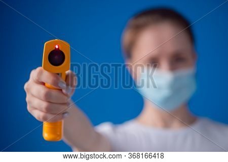 Woman In Medical Face Mask Holding Yellow Pyrometer To Measure Temperature Toward Camera - Close Up 