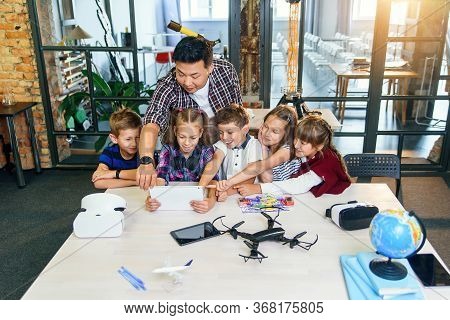 School Teacher At Desk Works With Five Young Pupils Using Digital Tablet Computer In Technology Clas