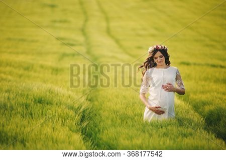 Young Pregnant Woman In Fieid Of Green Grass. Young Beautiful Pregnant Girl With A Wreath On Her Hea