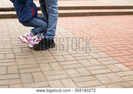 Dad Teaches His Little Daughter To Ride Four-wheeled Roller Skates. The Father Supports The Child By