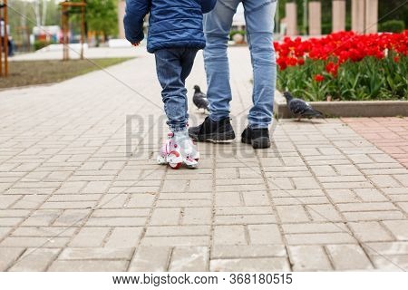 Dad Teaches His Little Daughter To Ride Four-wheeled Roller Skates. The Father Supports The Child By