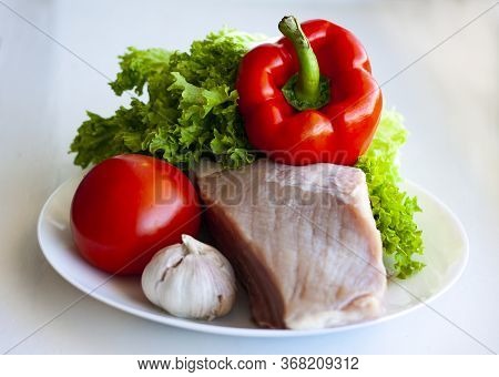 A Piece Of Raw Pork On A White Plate With Lettuce, Sweet Pepper, Tomato And Garlic. Healthy Eating A