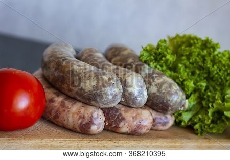 Raw Homemade Meat Sausages With Spices That Lie On A Wooden Board Near Tomato And Lettuce