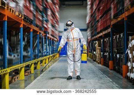 Man Worker With Protective Mask And Suit Disinfecting Industrial Factory With Spray Gun.