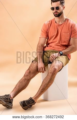 Young Man In Shorts, Summer T-shirt And Sunglasses Sitting On White Cube On Beige