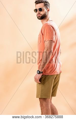 Handsome Young Man Posing In Shorts, Summer T-shirt And Sunglasses On Beige