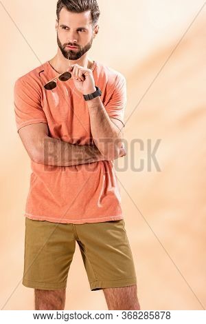 Handsome Man Posing In Shorts And Summer T-shirt Holding Sunglasses On Beige