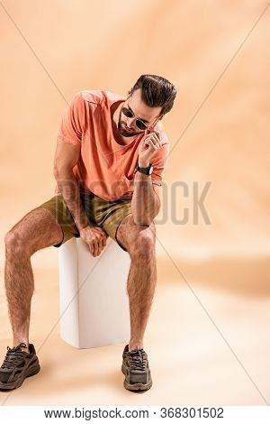 Fashionable Young Man In Shorts, Summer T-shirt And Sunglasses Sitting On White Cube On Beige