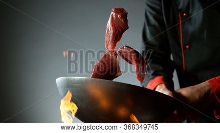 Closeup of chef throwing raw beef steaks from wok pan in fire. Fresh asian food preparation on dark background.