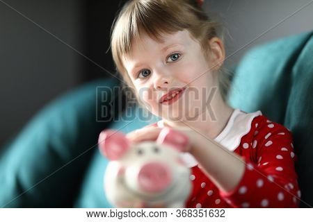 Portrait Of Smiling Beautiful Child Holding Piggy Bank Sitting On Sofa At Home. Lovely Blonde Kid Sa