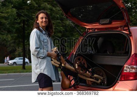 A Young Woman In Casual Clothes Takes A Folding Bike Out Of The Trunk Of A Car. Lays In The Trunk Of