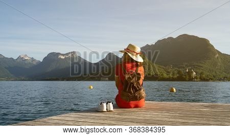 Girl With A Stalk Hat On And Red Dress Looking At The Mountains With Her Shoes On A Dock By A Lake. 