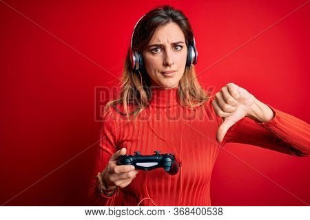 Young beautiful brunette gamer woman playing video game using joystic and headphones with angry face, negative sign showing dislike with thumbs down, rejection concept