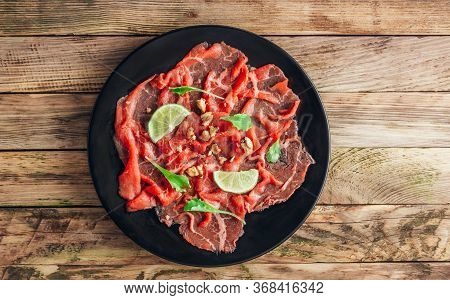 Black Plate With Carpaccio Of Beef With Baby Arugul And Lime On Rustic Wooden Background. Top View. 