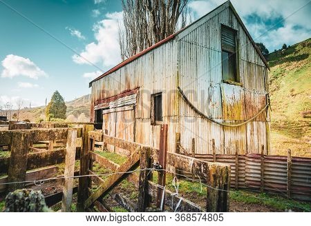 Old Rural Corrugated Iron Building And Stock Yard Railings In Shade With Sun On Surrounding Hills In